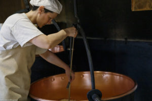 Une femme, fromagère, décaille le fromage à l'aide d'un tranche-caillé dans un chaudron en cuivre, à la ferme Chez Valentin à Foncine-le-Haut